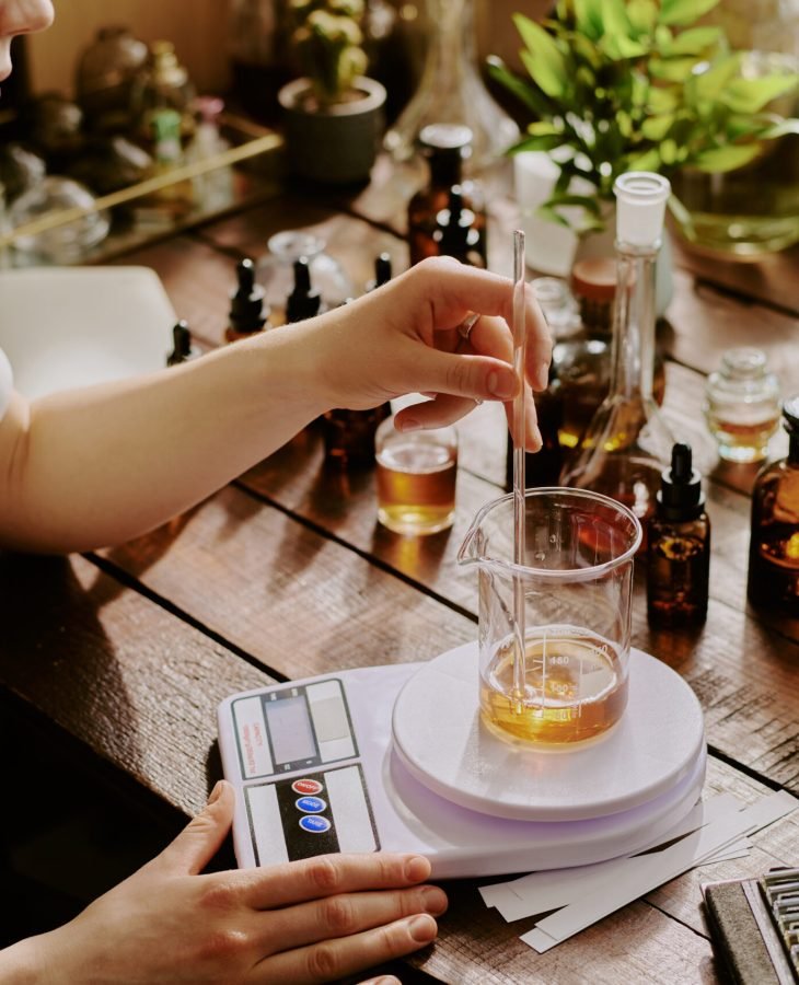 Young Perfumer Blending Essential Oils Young female perfumer blending essential oils while she weighing it with plastic kitchen scale